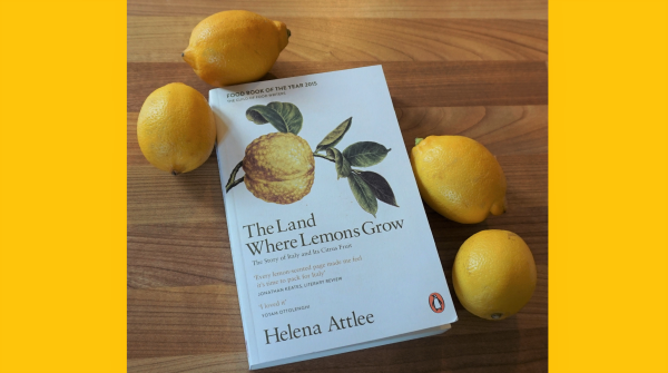 A book titled The Land Where Lemons Grow by Helena Attlee is placed on a wooden surface, surrounded by four whole lemons. The book cover features an illustration of a lemon and leaves.