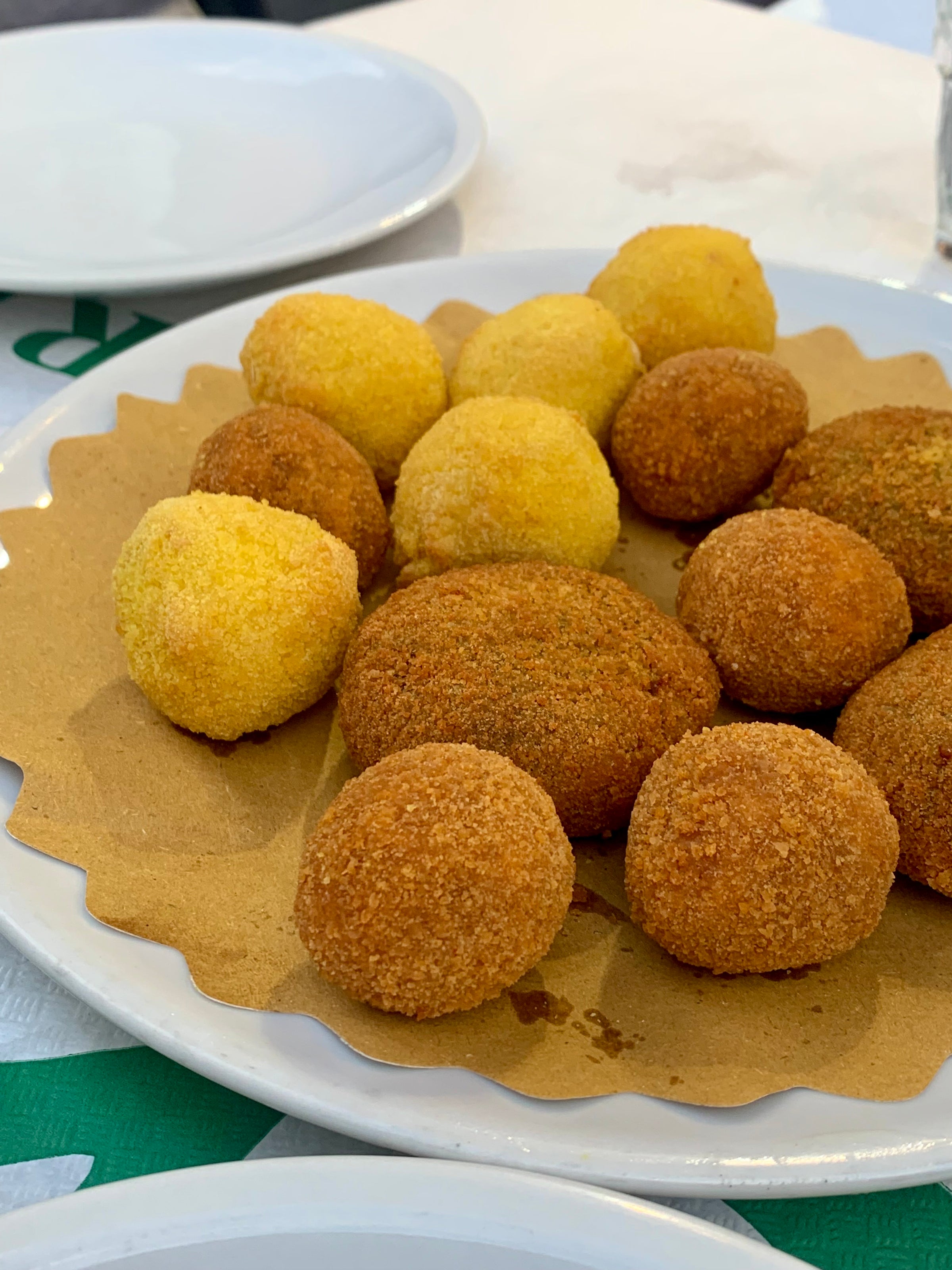 A plate with various round, golden-brown and yellow fried snacks, placed on brown paper atop a white plate. An empty white plate is visible in the background.