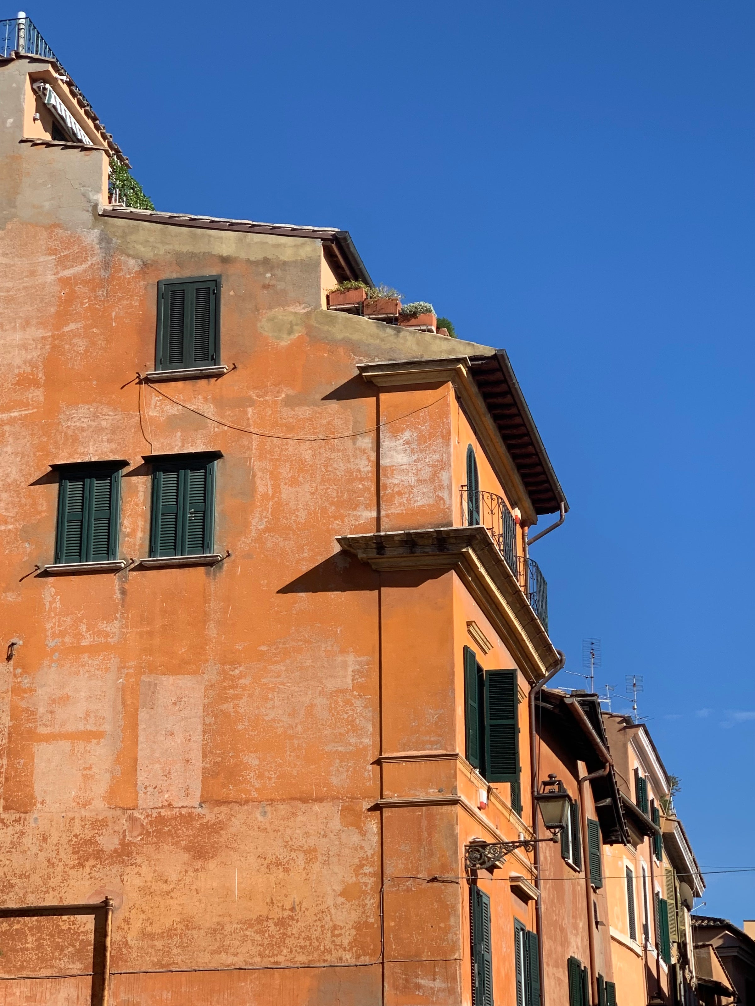 An orange, weathered building with green shutters stands under a clear blue sky. The sunlight casts sharp shadows on the facade, highlighting architectural details and a small balcony with potted plants.