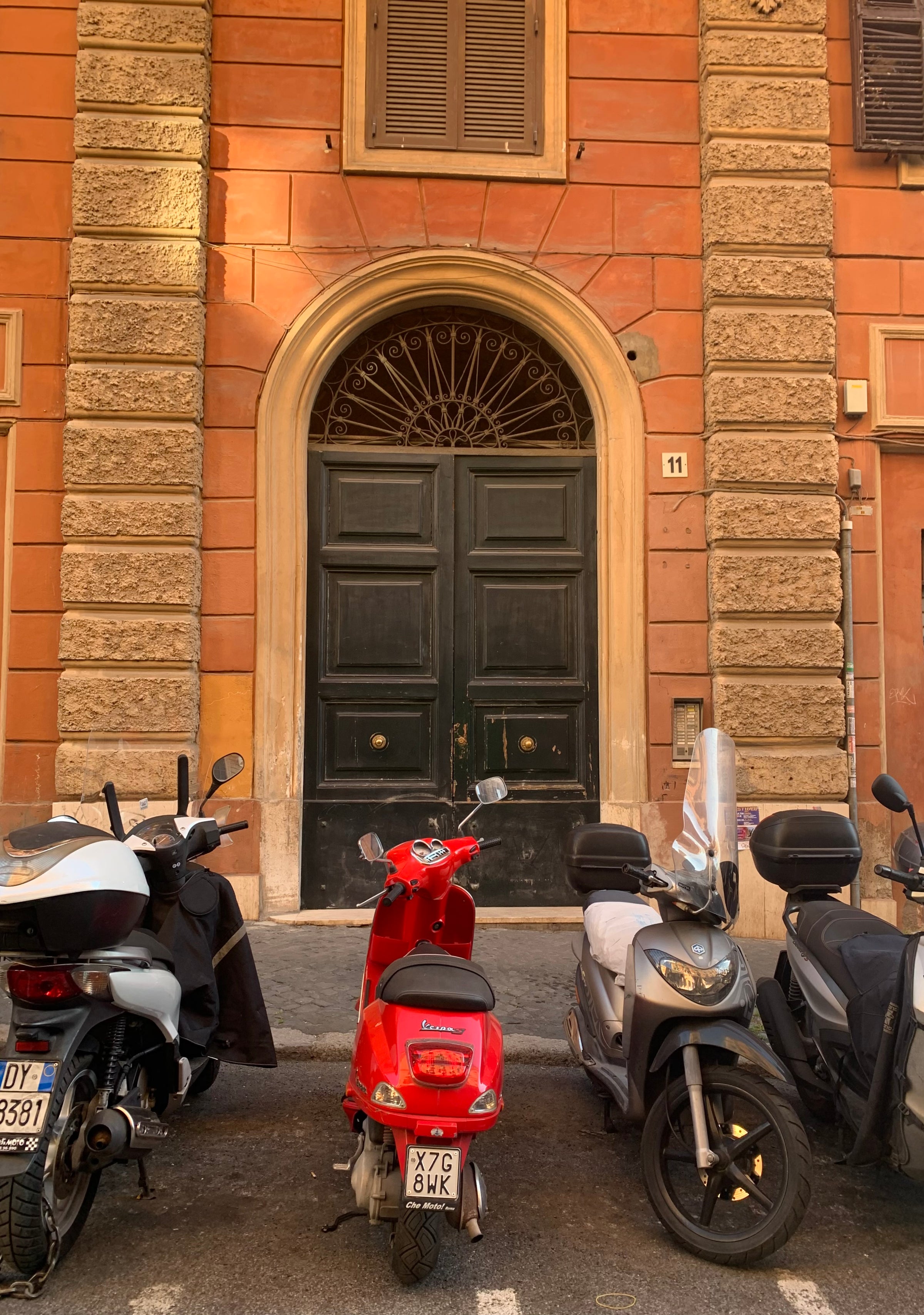 Four scooters, including a bright red one in the center, are parked in front of a large, ornate dark green double door set in an orange brick building with a decorative arch and the number 11 above the entrance.