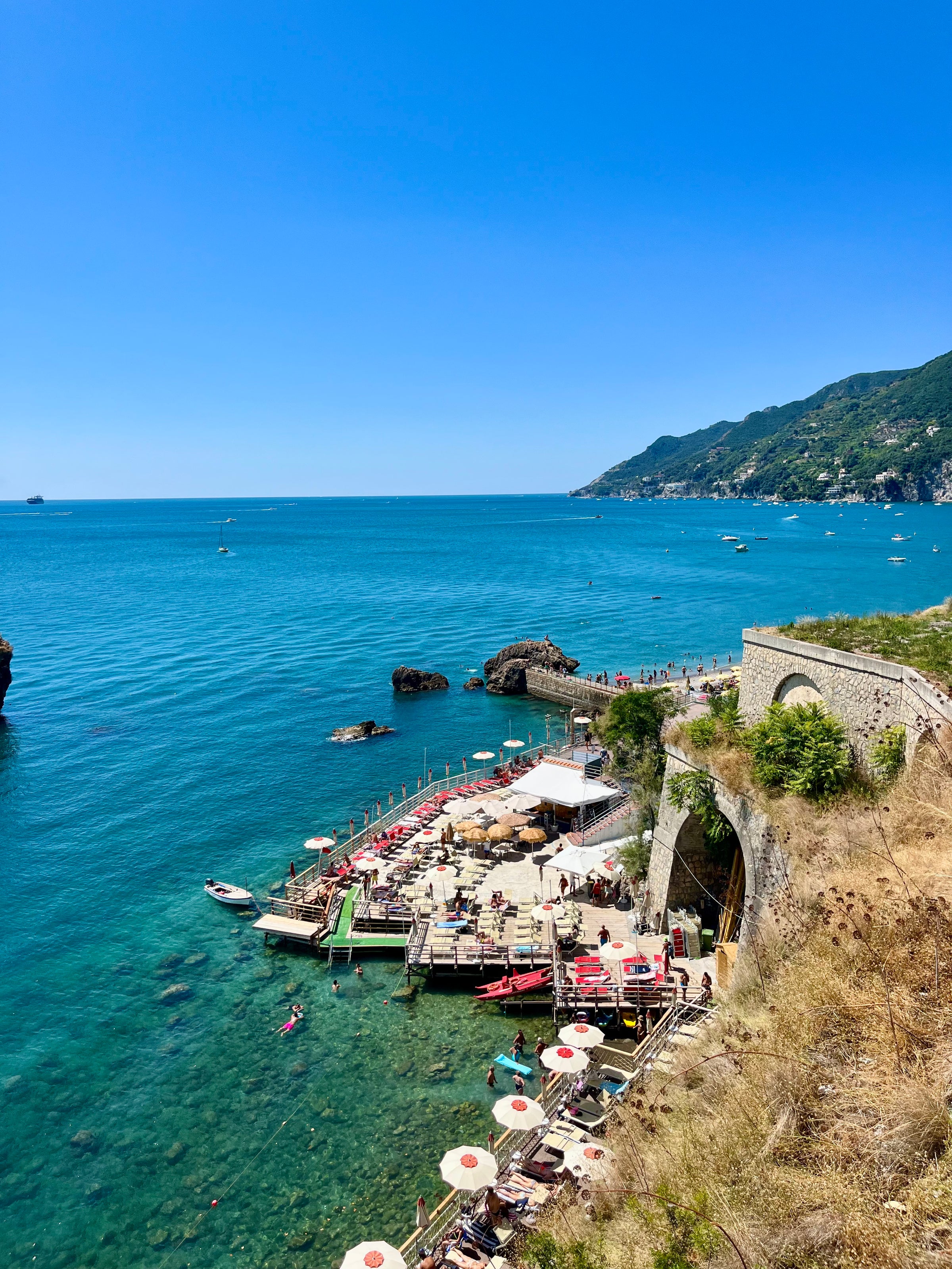 A vibrant seaside view shows a beach club with sunbeds and umbrellas on wooden decks over turquoise water, people swimming, and a curving stone road along a hillside with lush greenery and distant mountains under a clear blue sky.