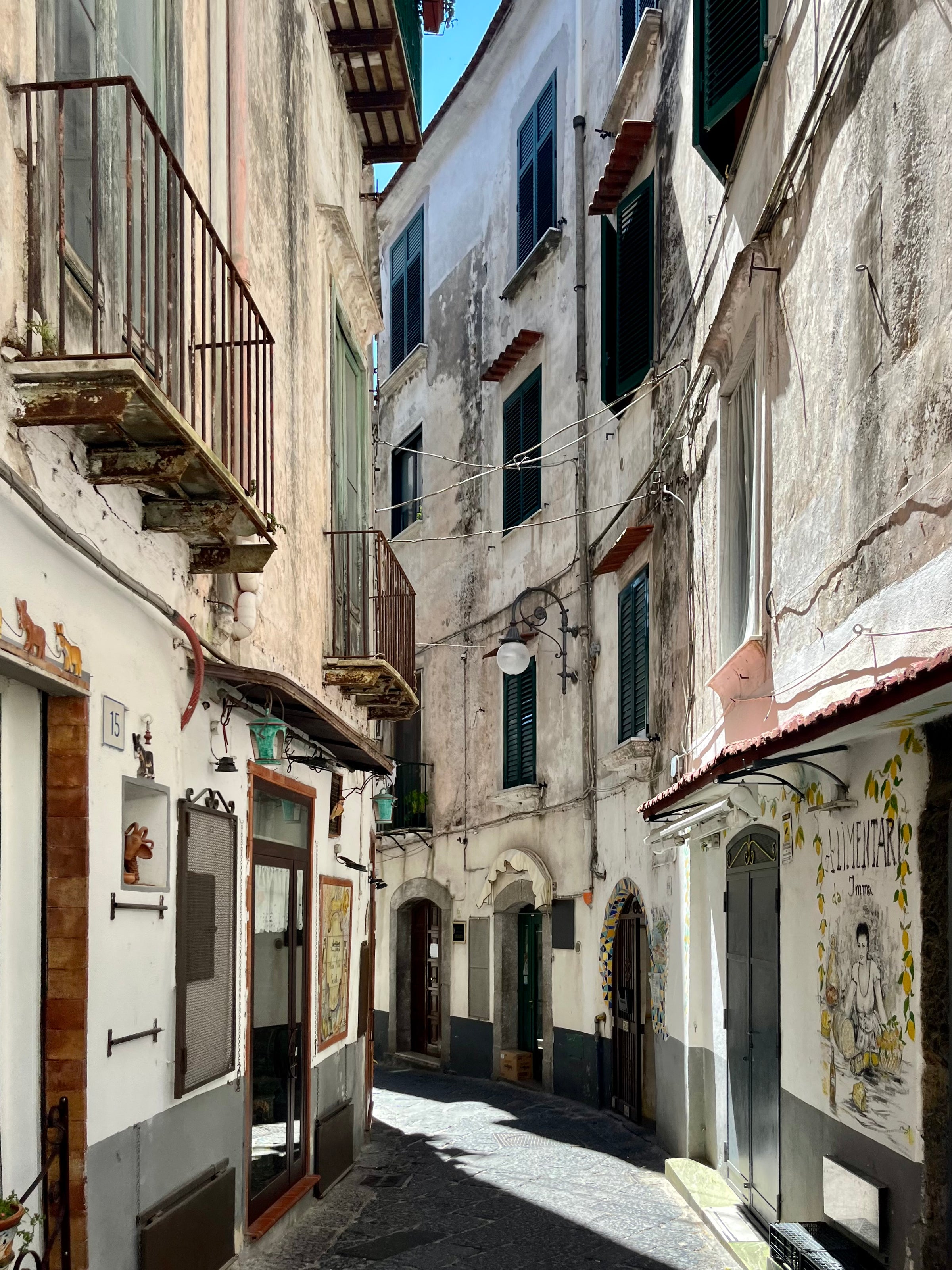 A narrow, sunlit alleyway in an old European town, lined with weathered buildings, shuttered windows, small balconies, and colorful wall art. Shadows and sunlight highlight the texture of the historic architecture.