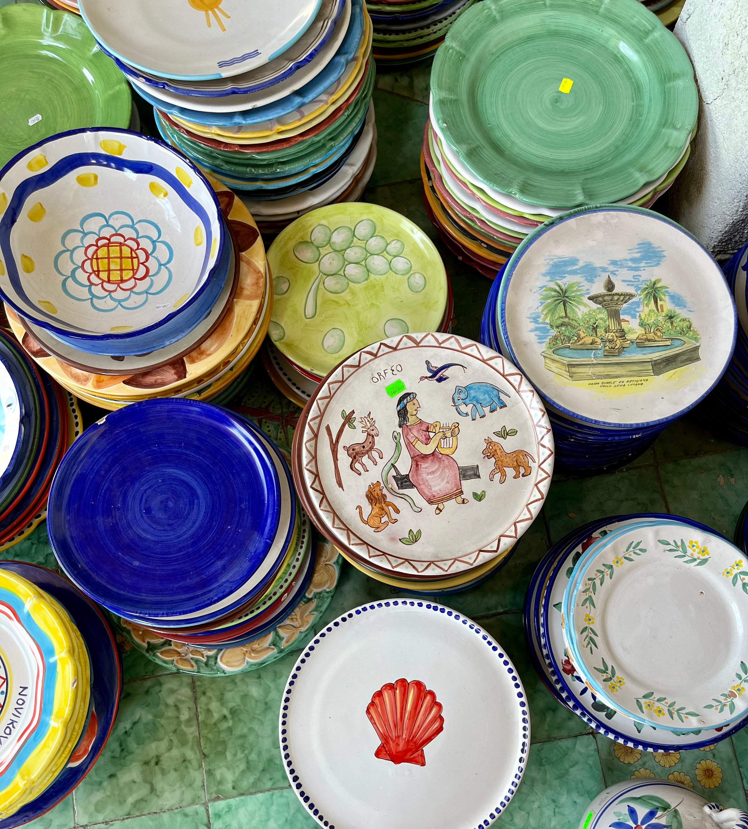 Stacks of colorful ceramic plates with various painted designs, including fruits, flowers, animals, and scenic landscapes, are arranged on a green tiled floor. The plates feature bright colors and intricate patterns.
