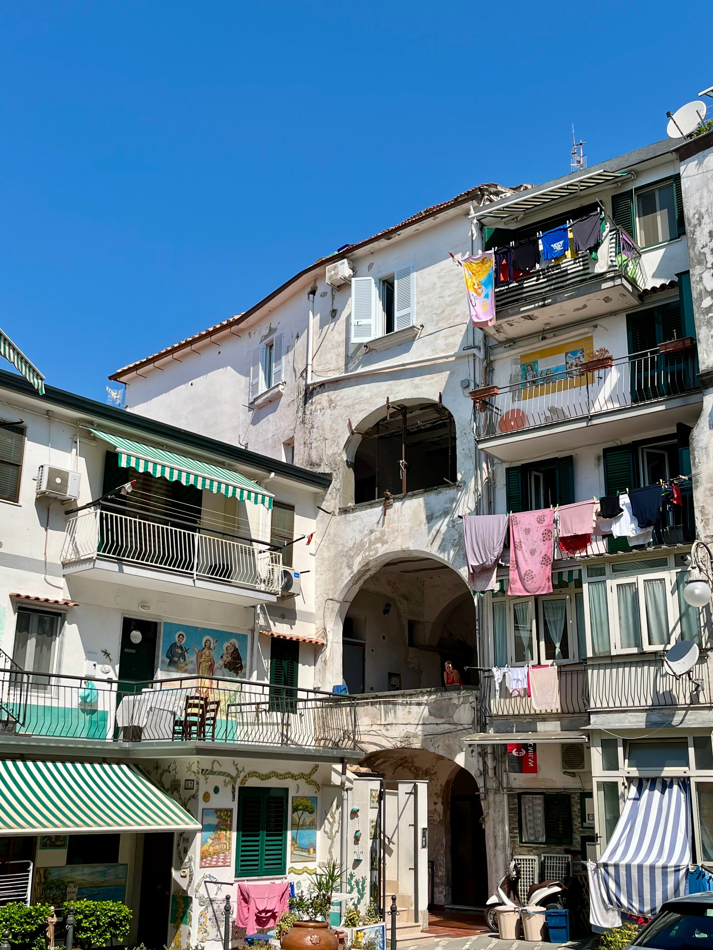 Colorful apartment buildings with balconies display laundry hanging out to dry under a clear blue sky. Green shutters, striped awnings, and small murals add charm to the sunny Mediterranean street scene.