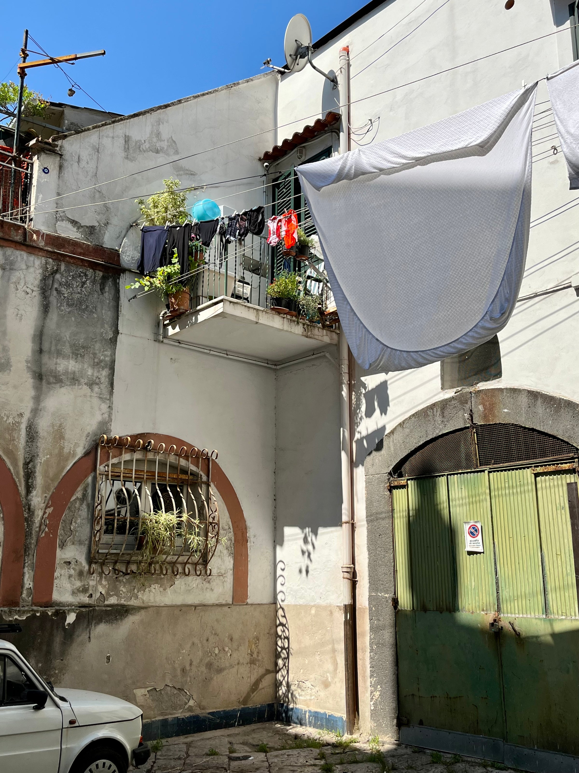 A sunny courtyard with a small balcony displaying hanging laundry, potted plants, and an umbrella. A large white sheet dries overhead. Below, an old car is parked next to a green metal gate and a barred window.