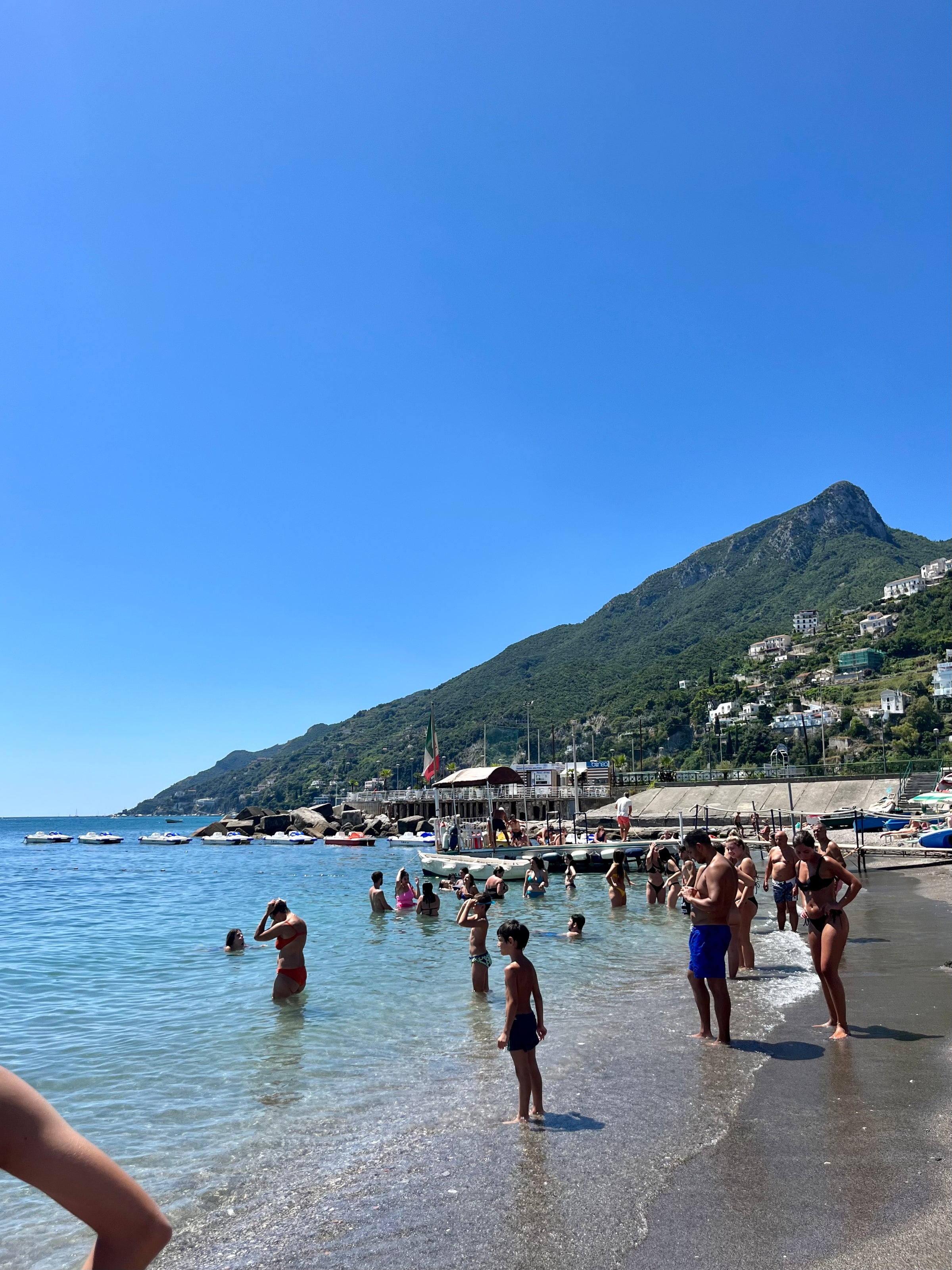 People enjoy a sunny day at a beach with clear blue water, mountains and green hills in the background, and boats docked near the shore. Swimmers and sunbathers are scattered along the sand and in the shallow water.