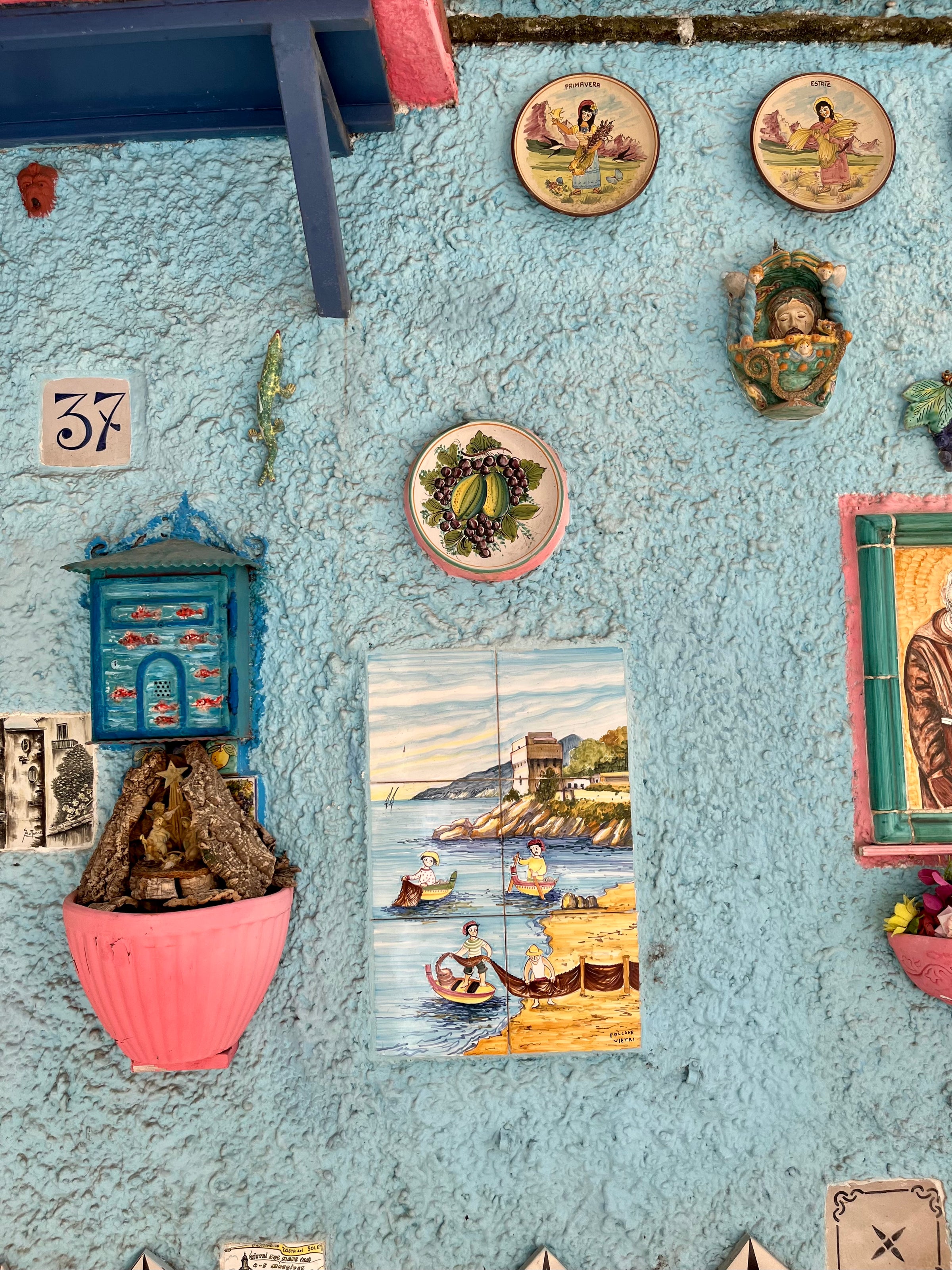 A textured blue wall decorated with colorful plates, religious icons, a wall fountain, a small blue shrine, and ceramic figures. A painted tile mural shows boats on water near a coastal town.