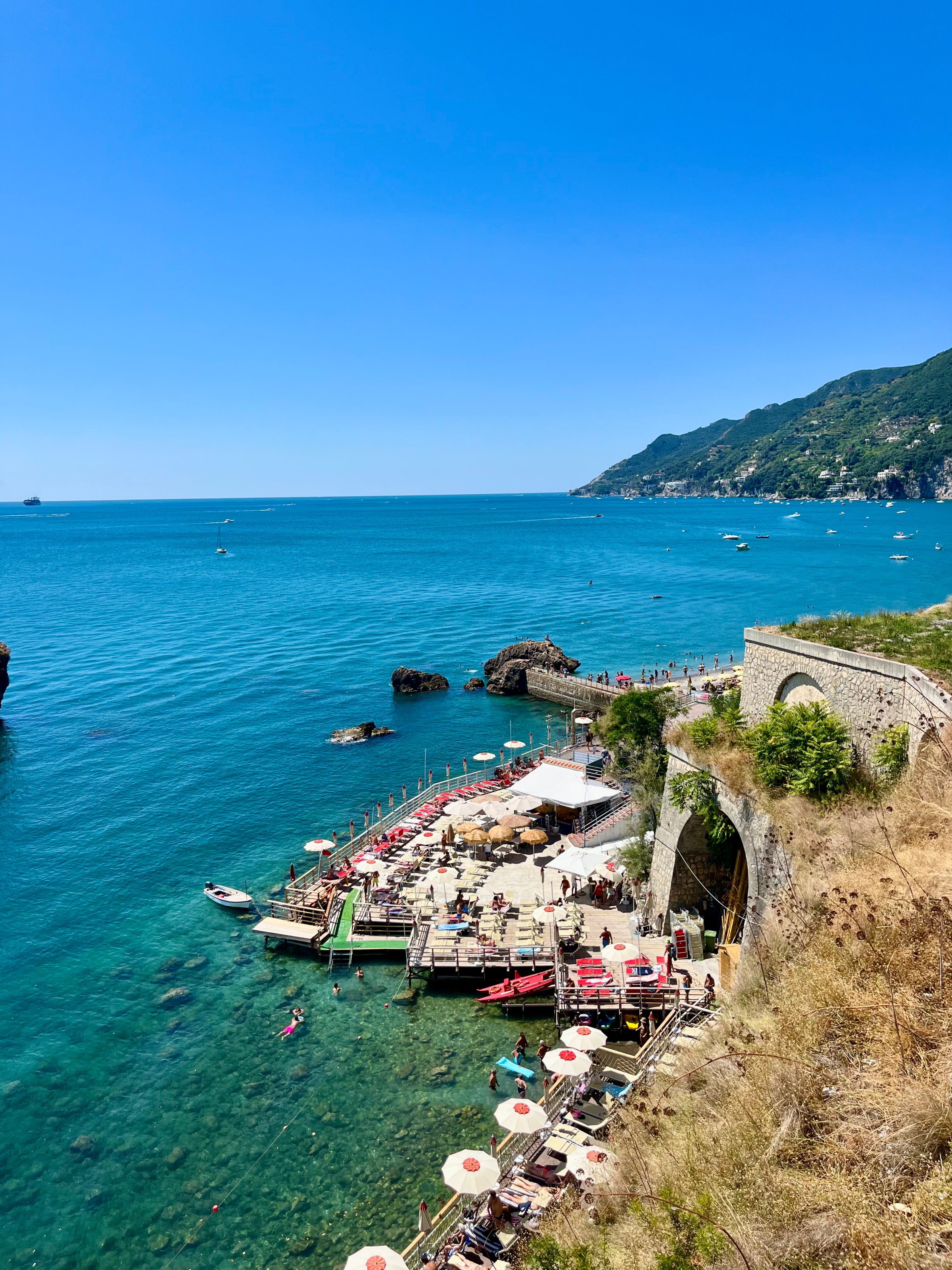 A seaside view of a rocky coastline with a beach club featuring sunbeds, umbrellas, and people relaxing on a wooden platform over clear blue water, surrounded by lush green hills under a clear blue sky.