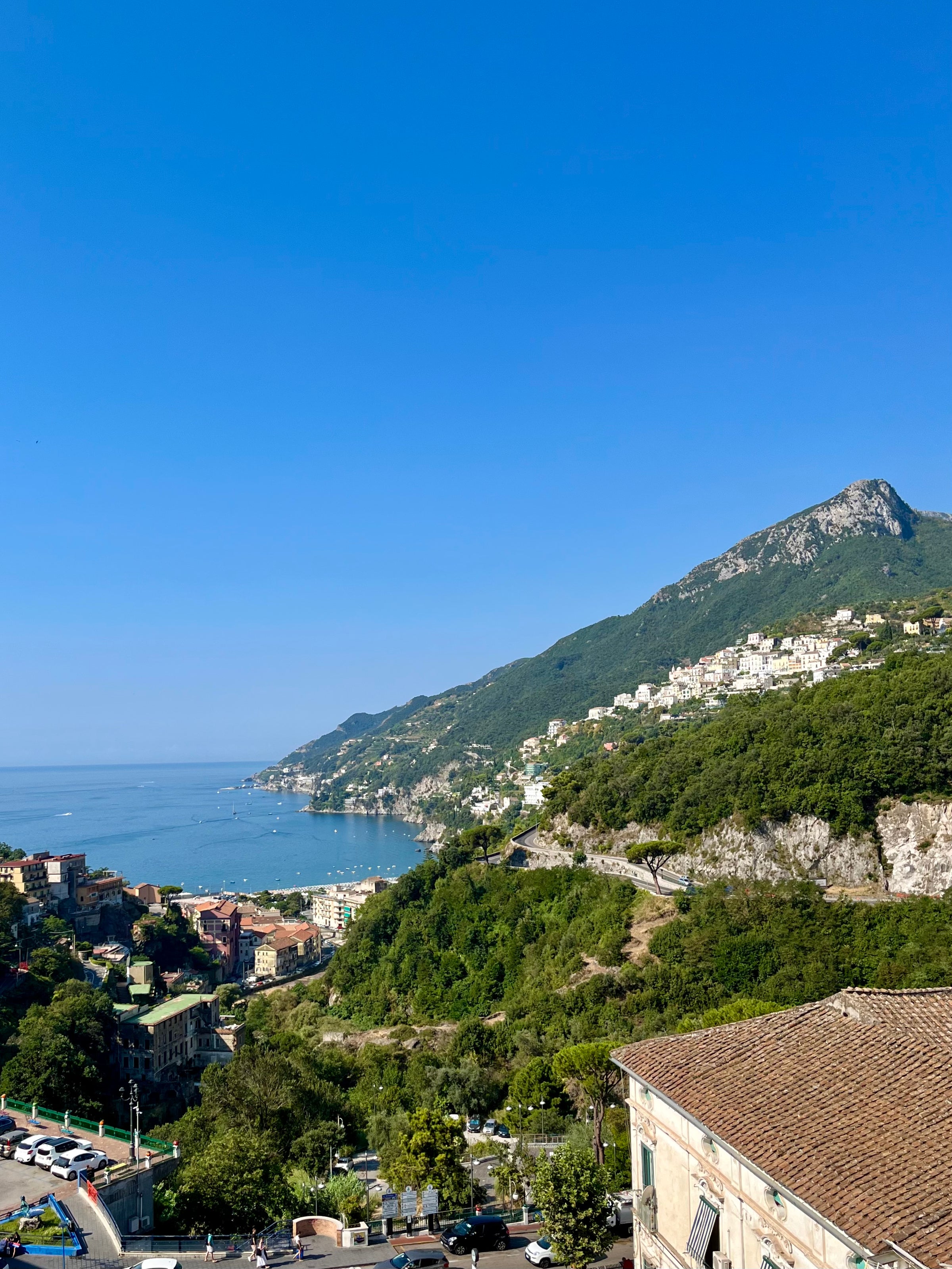 View of a coastal town with green hills, scattered buildings, a curving shoreline, and clear blue sky, with the sea stretching into the distance and a mountain rising on the right.