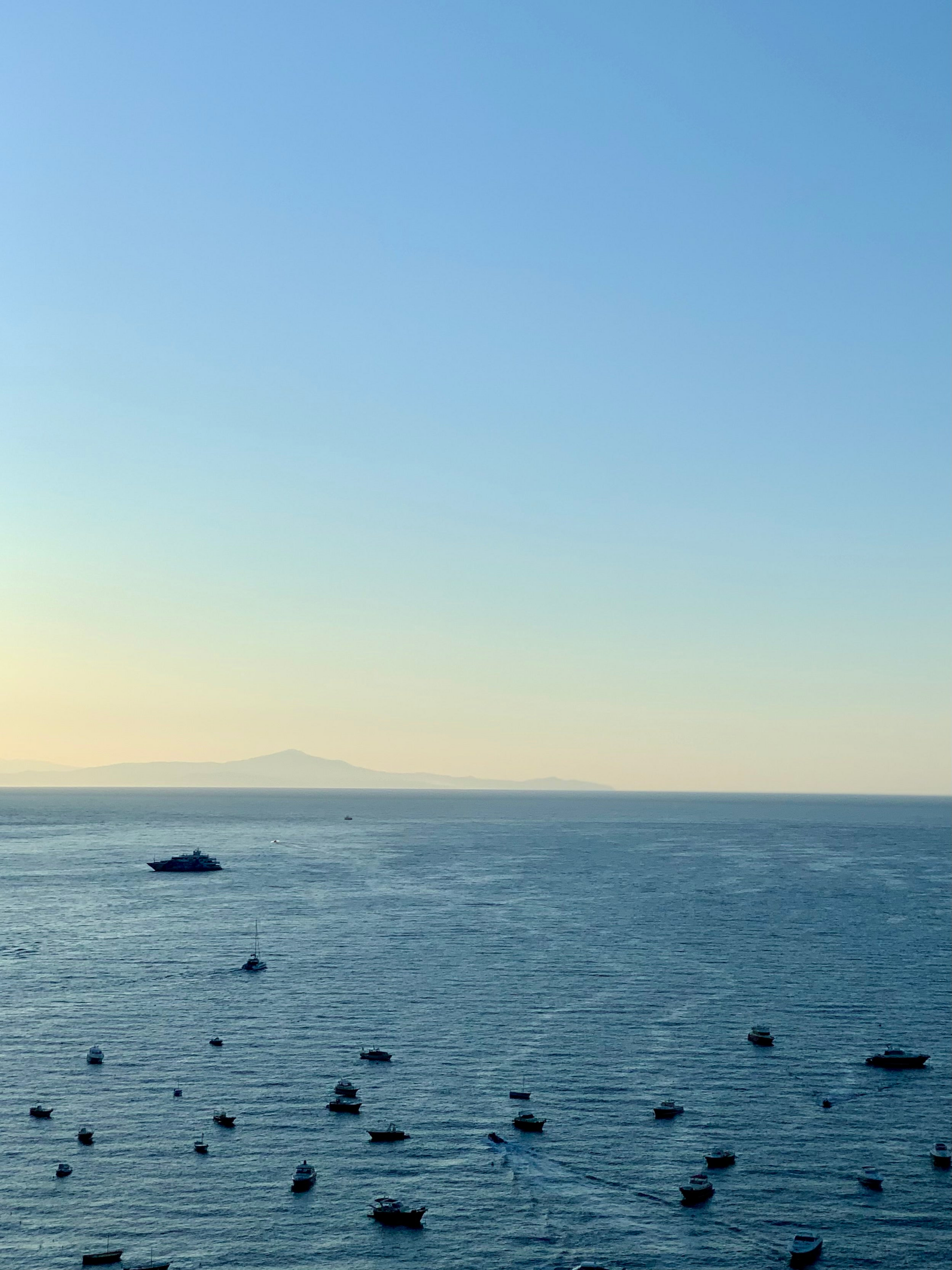 A calm sea with many small boats scattered across the water under a clear blue sky at sunset, with distant mountains visible on the horizon.