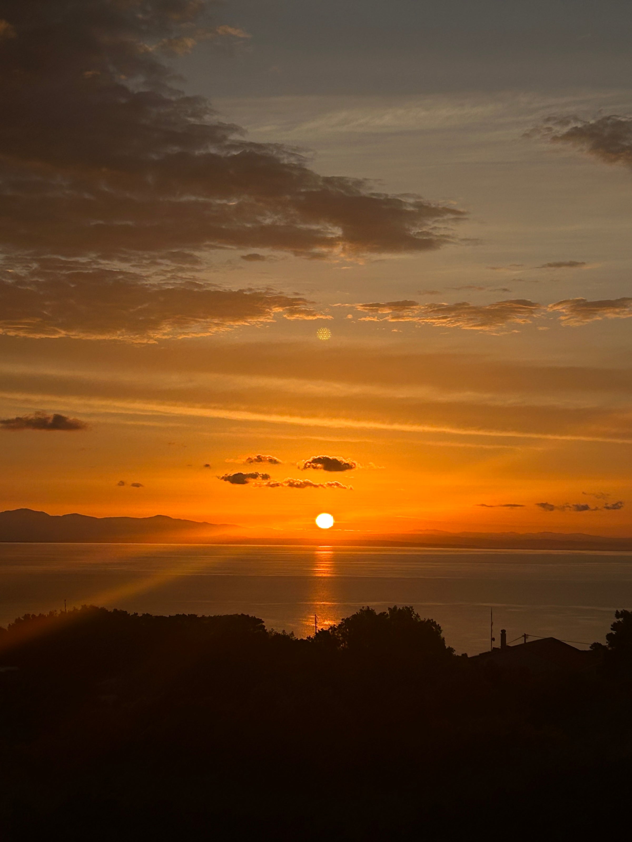 A bright orange sun sets over a calm body of water, casting golden reflections. Silhouetted trees and rooftops are visible in the foreground, with mountains and scattered clouds in the background.