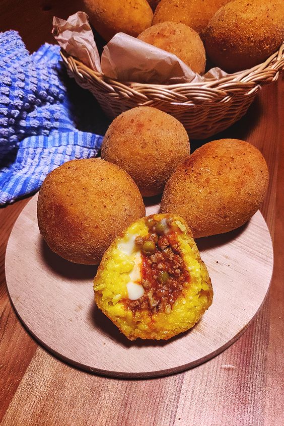 A wooden plate holds several arancini, with one cut open to reveal a filling of rice, meat, peas, and melted cheese. More arancini are in a basket in the background next to a blue-striped cloth.