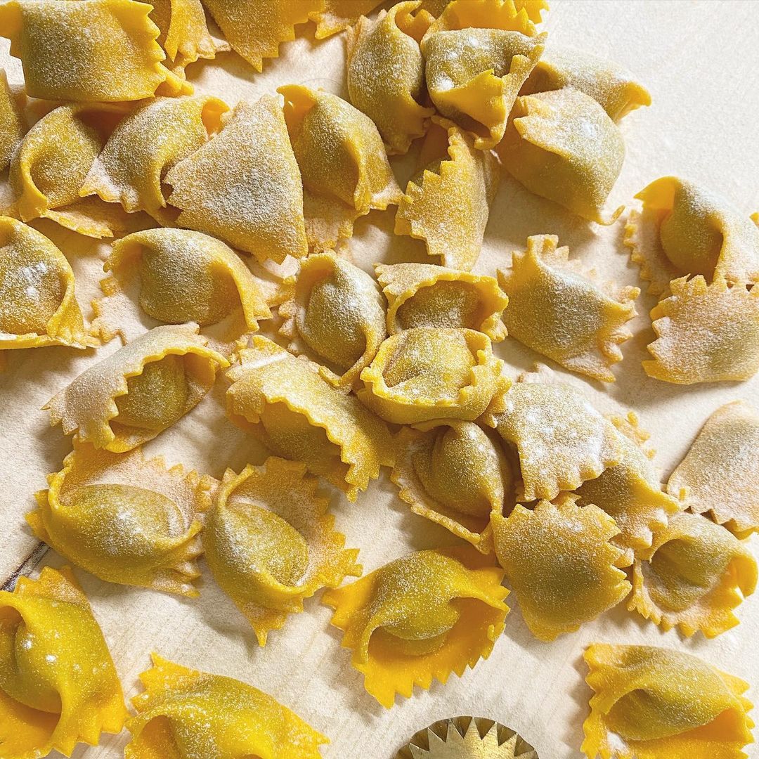Several pieces of uncooked, yellow tortellini pasta are scattered on a floured surface, showcasing their folded and crimped edges. A round pasta cutter is partially visible at the bottom.