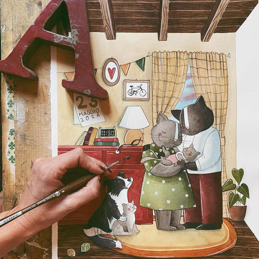 A hand draws a cozy scene of two cats in clothes hugging with their kittens, a dog, and a kitten nearby in a warmly decorated room. A large letter A and a calendar reading 23 Maggio 2024 are visible.