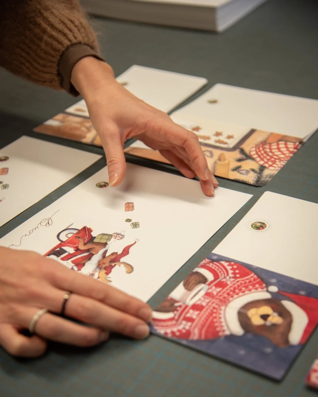 A person arranges illustrated holiday cards on a table. The cards feature festive images, including Santa Claus and animals in winter clothing. The persons hands are visible, and theyre wearing a brown sweater.