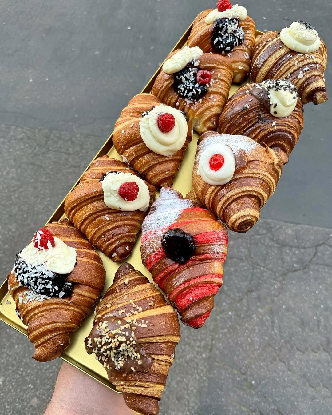 A hand holds a tray filled with assorted croissants, each topped with different creams, fruits, chocolate, and nuts, displayed on a golden rectangular tray against a gray pavement background.