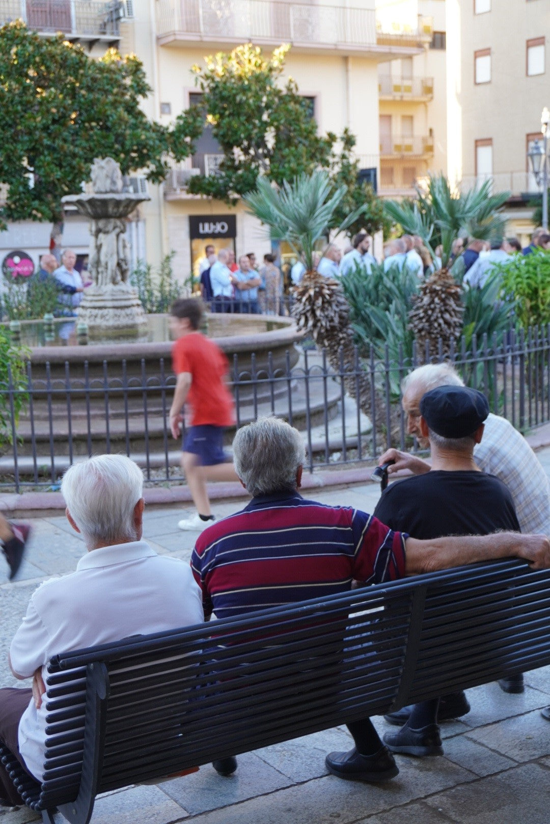 Three elderly men sit on a bench facing a fountain in a lively town square, while people walk and gather in the background among trees and buildings. A child runs past them, creating a sense of movement.