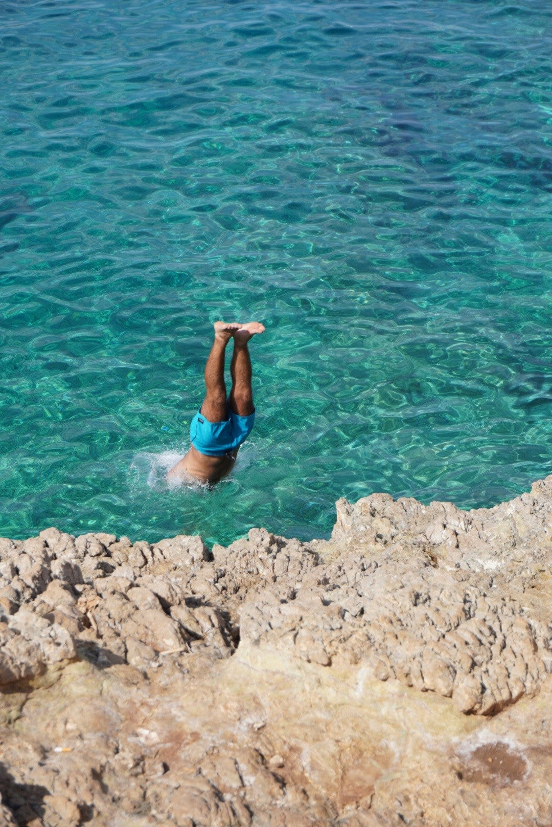 A person in blue swim trunks is diving headfirst into clear turquoise water from a rocky ledge, with only their legs and feet visible above the surface.