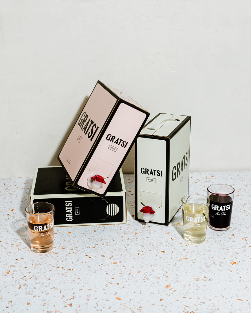 Three boxed wines labeled GRATSI in pink, white, and black are stacked on a terrazzo surface. Three glasses with GRATSI printed on them hold rosé, white, and red wine in front of the boxes.
