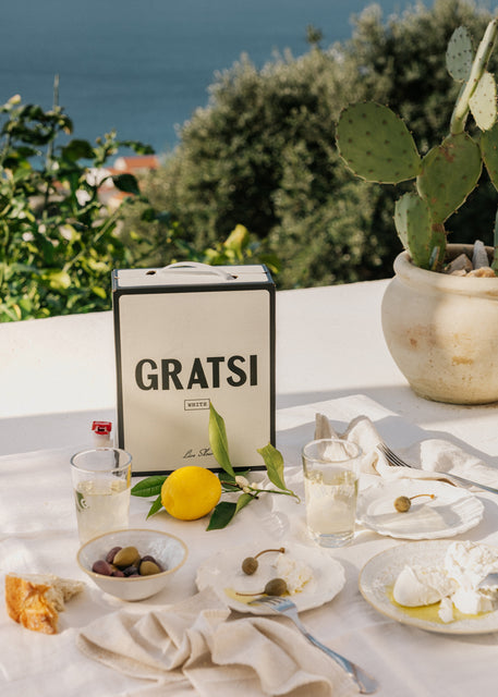 Outdoors, a picnic table is set with plates of cheese, olives, and bread beside a “Gratsi” Bundle & Save box and a lemon with leaves. Behind are a potted cactus, greenery, and a sea view.