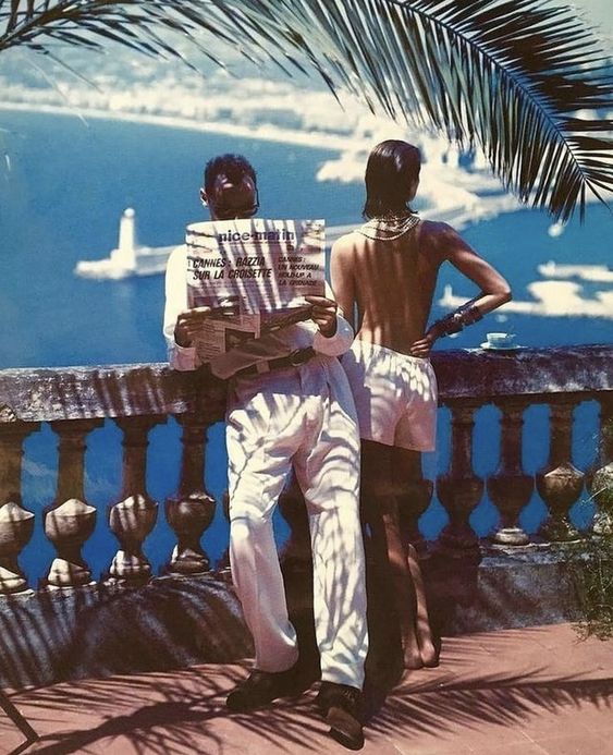 A man in white clothes reads a newspaper on a sunny terrace overlooking a harbor, while a woman in a backless dress stands beside him, both partly shaded by palm fronds. Sailboats and water are visible in the background.