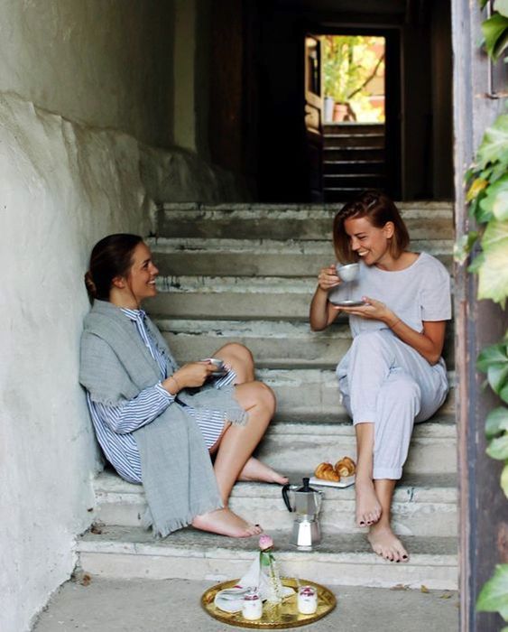 Two women sit barefoot on stone steps, enjoying coffee and pastries together. One wears a striped dress with a shawl, the other light pajamas. They smile and chat, surrounded by a cozy, rustic atmosphere.