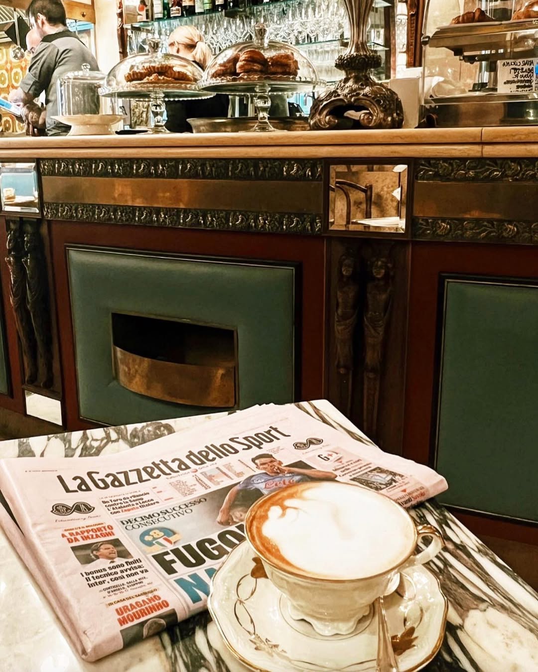 A marble café table with a cappuccino in a white cup and saucer, and a folded Italian newspaper, La Gazzetta dello Sport. Behind, a vintage-style counter with pastries, glasses, and a barista working.