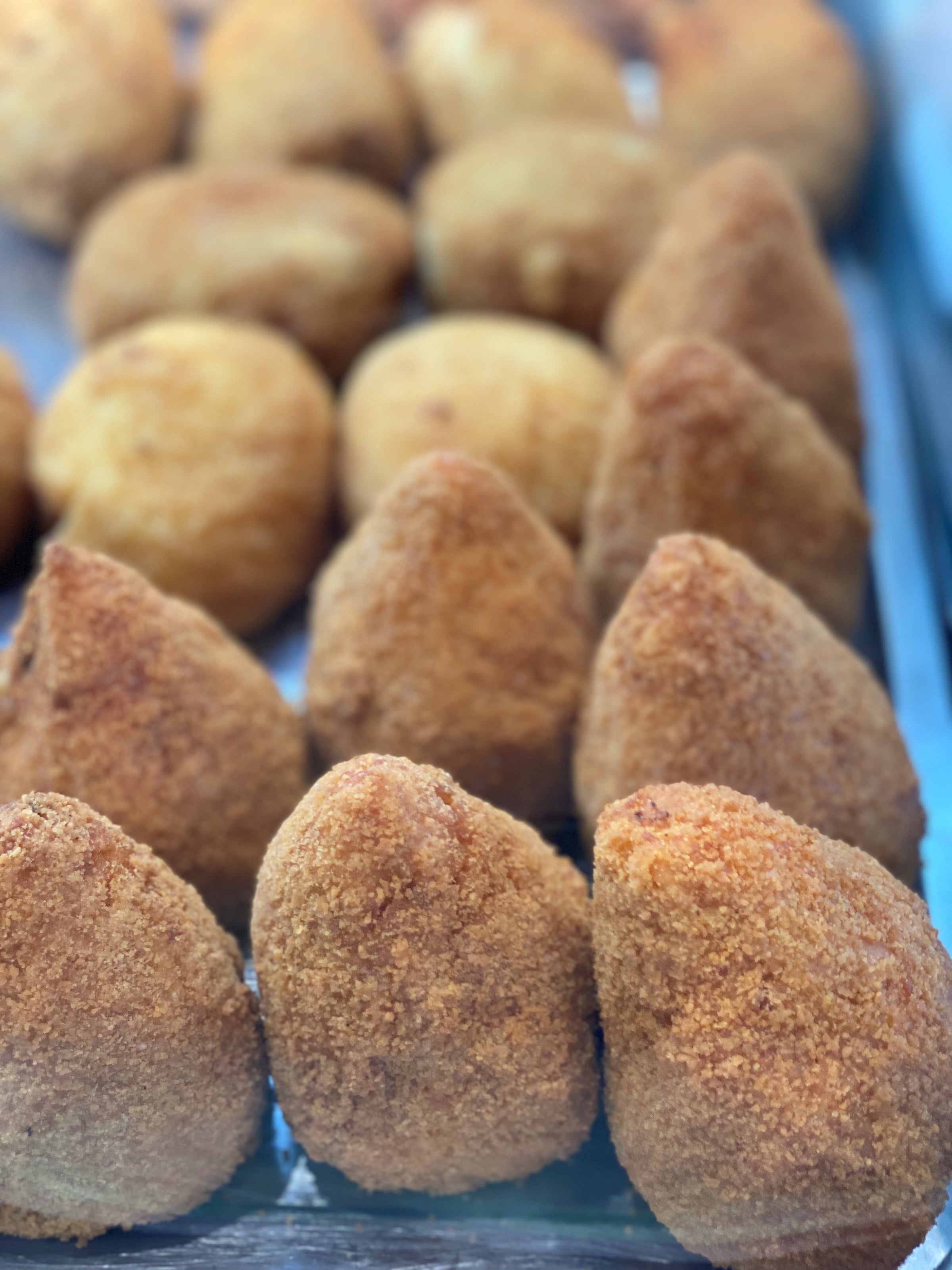 A close-up of rows of golden-brown, breaded and fried snacks with a conical shape, displayed on a metal tray. The snacks appear crispy and are arranged neatly in lines.
