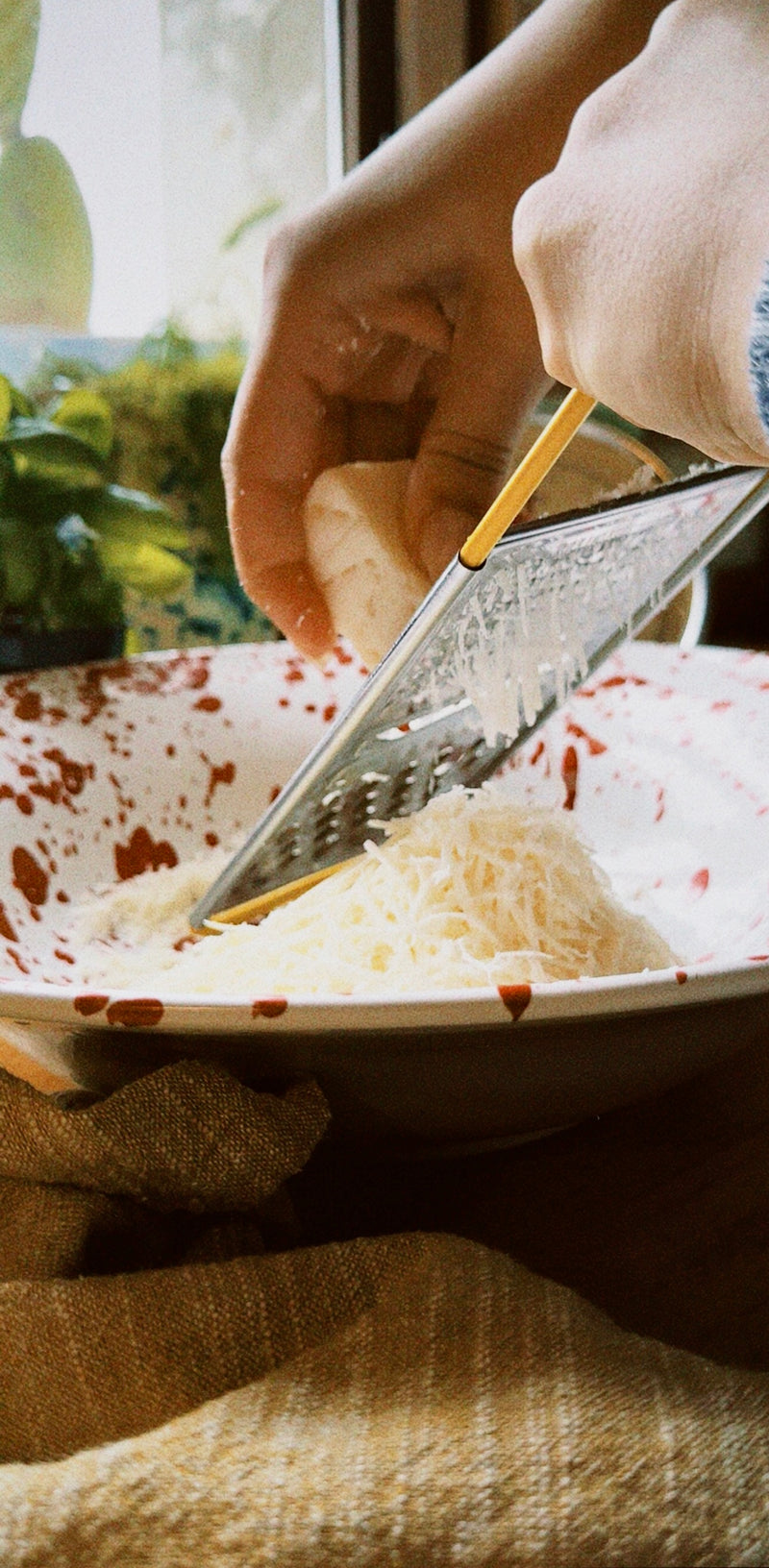 A person grates cheese into a white bowl with red spots, using a metal grater. Green plants are visible in the background near a window.