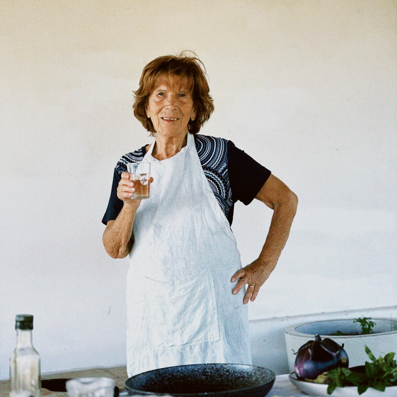 An elderly woman wearing a white apron stands in a kitchen, smiling and holding a glass. There are cooking ingredients, including a bottle of oil and a bowl with an eggplant, on the counter in front of her.