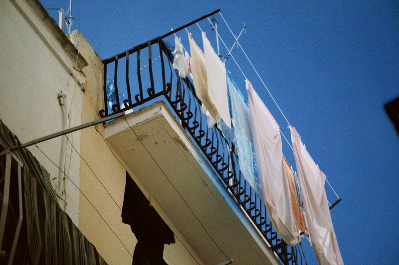 Clothes hang on a line above a small balcony with black railings, attached to a light-colored building, set against a clear blue sky.