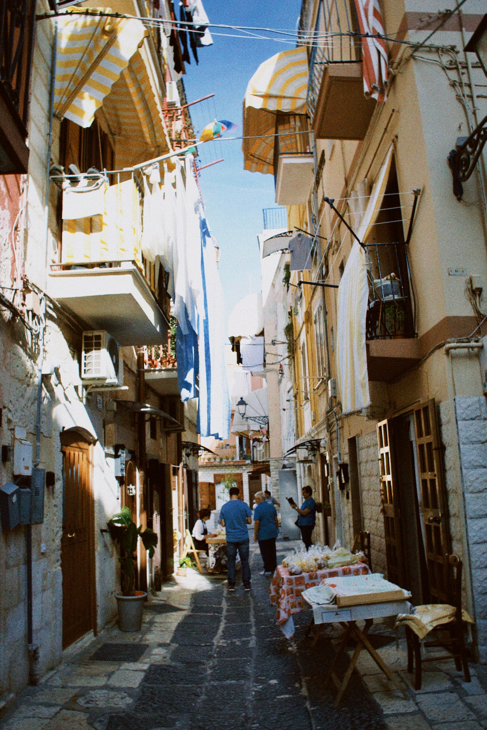 A narrow, sunlit alleyway with colorful laundry hanging above, striped awnings, and tables set along the cobblestone street. Several people stand and walk, creating a lively, inviting atmosphere.