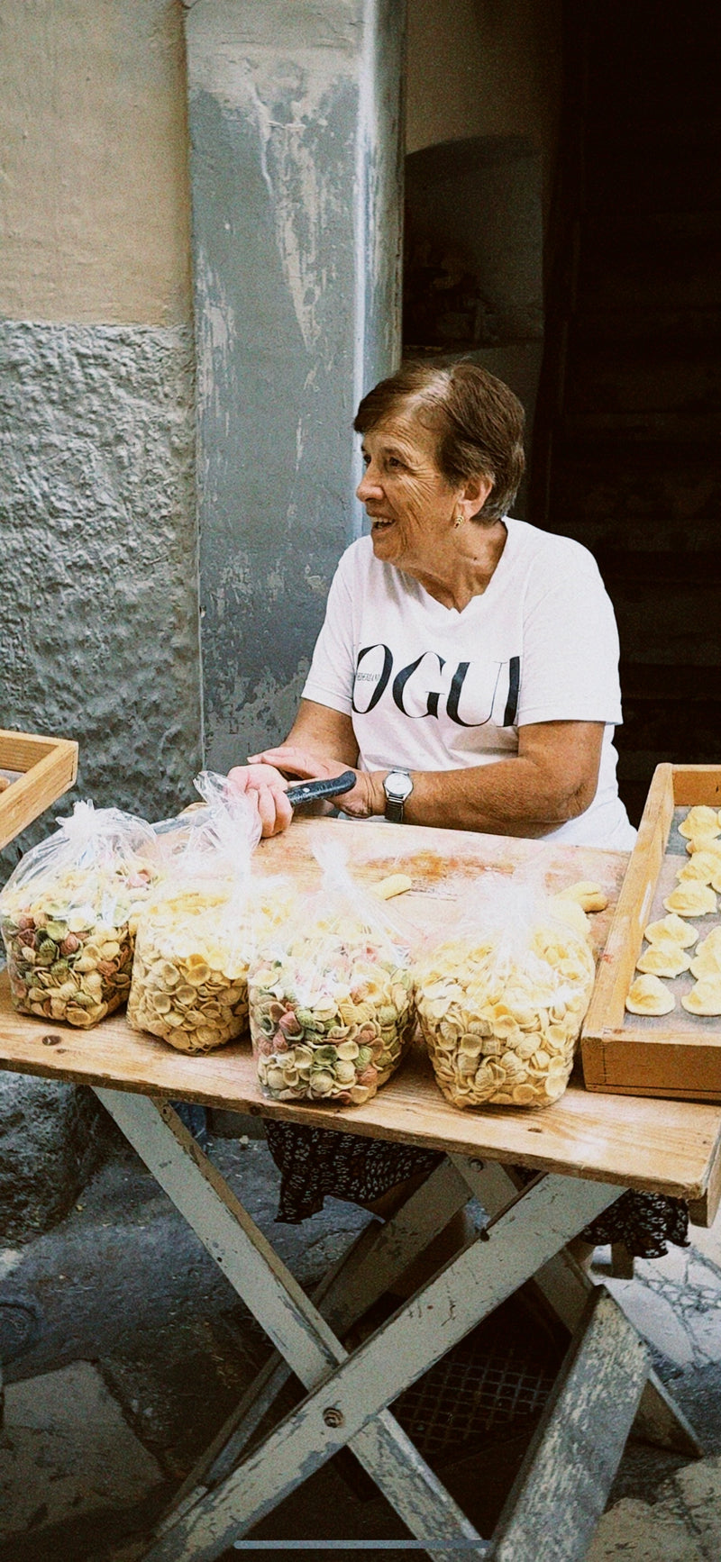 An elderly woman with short hair, wearing a white VOGUE t-shirt, sits at an outdoor table selling homemade pasta displayed in wooden trays and plastic bags. She is smiling and appears to be interacting with someone.