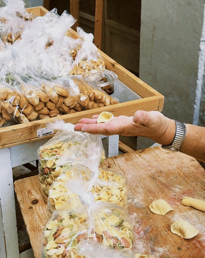 A hand holds a piece of fresh pasta over a wooden table with bags of various types of dried pasta and snacks displayed for sale.
