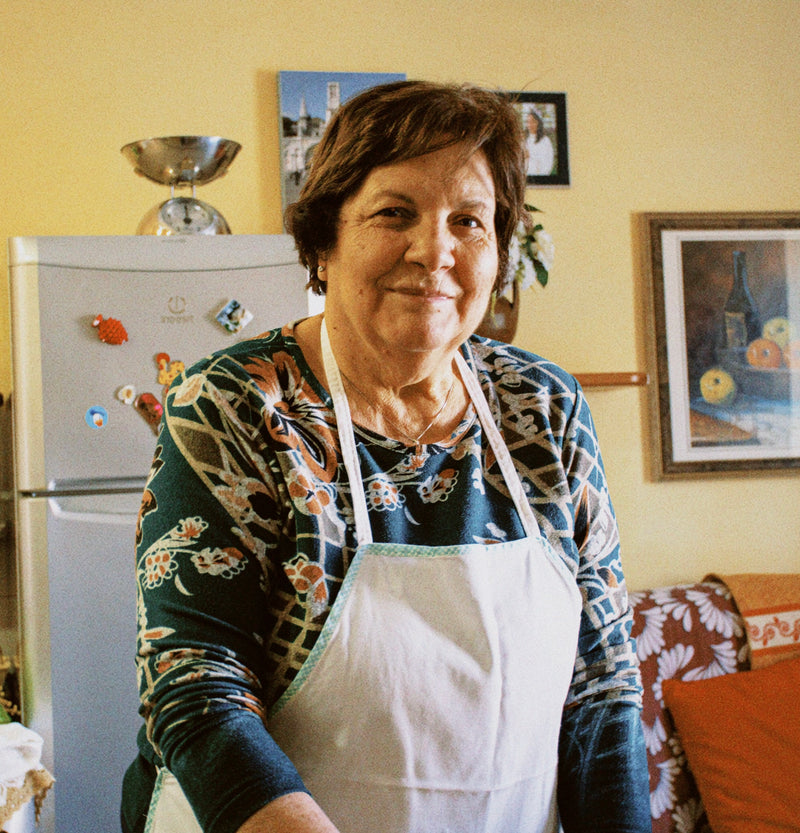 An older woman wearing a patterned shirt and a white apron stands in a cozy kitchen, smiling at the camera. There is a fridge with magnets, wall art, and a decorative scale in the background.