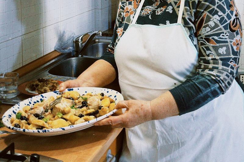 A person wearing a white apron and patterned shirt holds a large platter of homemade food in a kitchen, with a sink and countertop in the background. The dish appears to be a mix of pasta, vegetables, and olives.