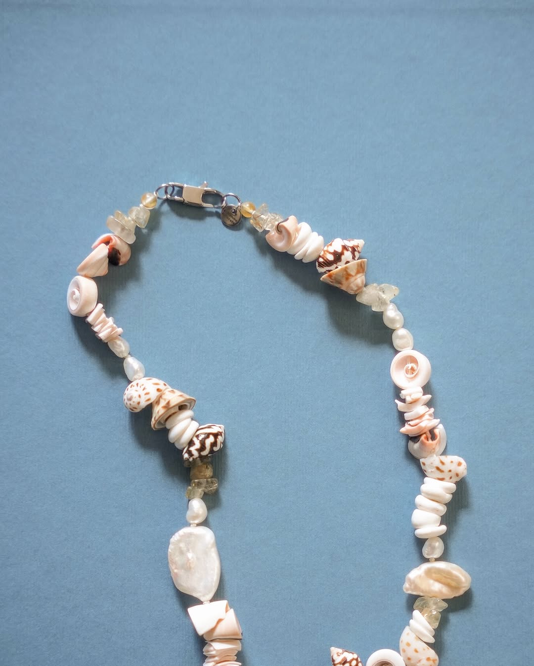 A necklace made of assorted seashells and translucent beads is arranged in a curve on a flat, light blue surface. The shells are various shapes and colors, adding texture and visual interest to the design.