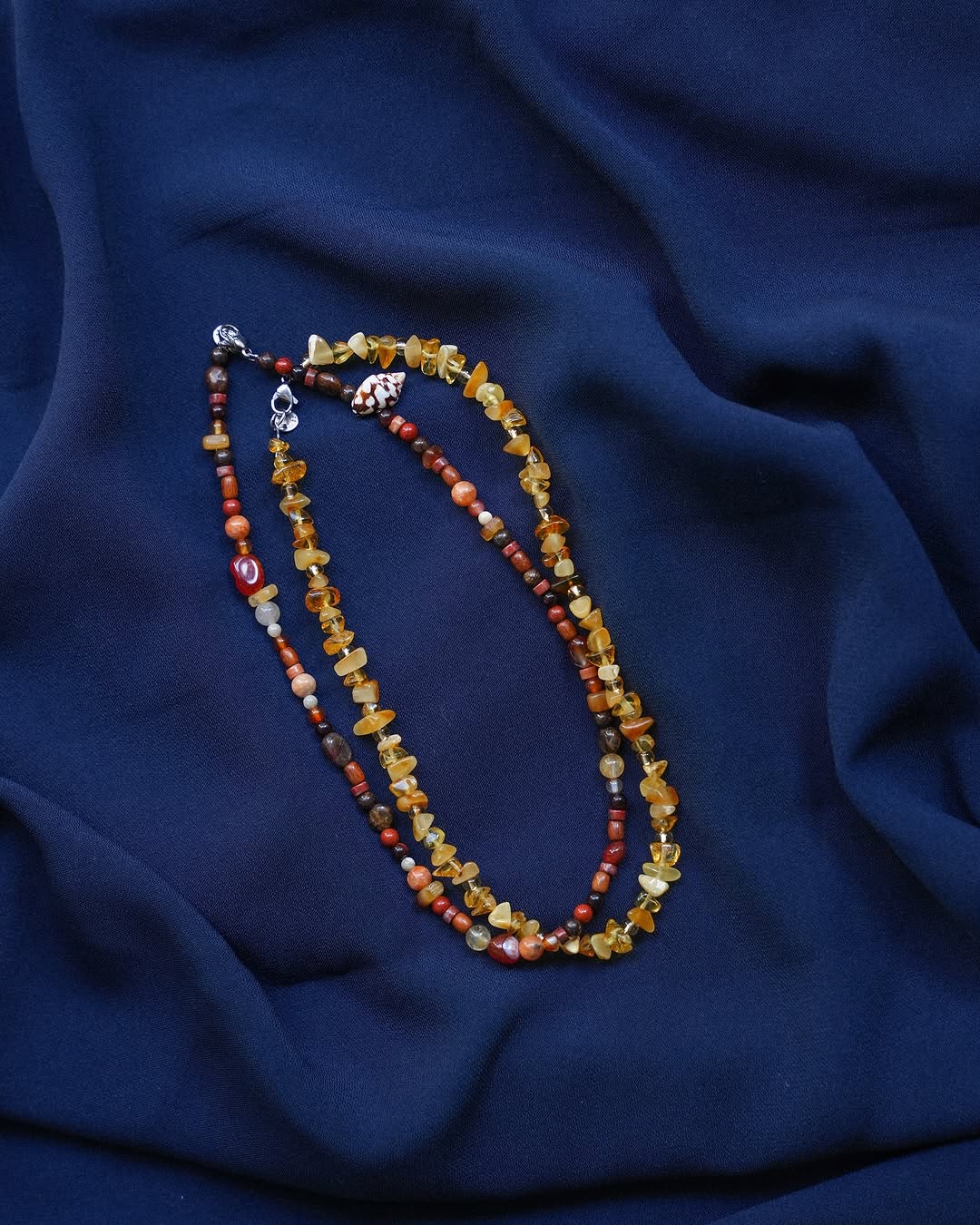 Two beaded necklaces with amber and brown stones are laid on a deep blue, softly draped fabric background. The necklaces feature irregularly shaped beads and metallic clasps.
