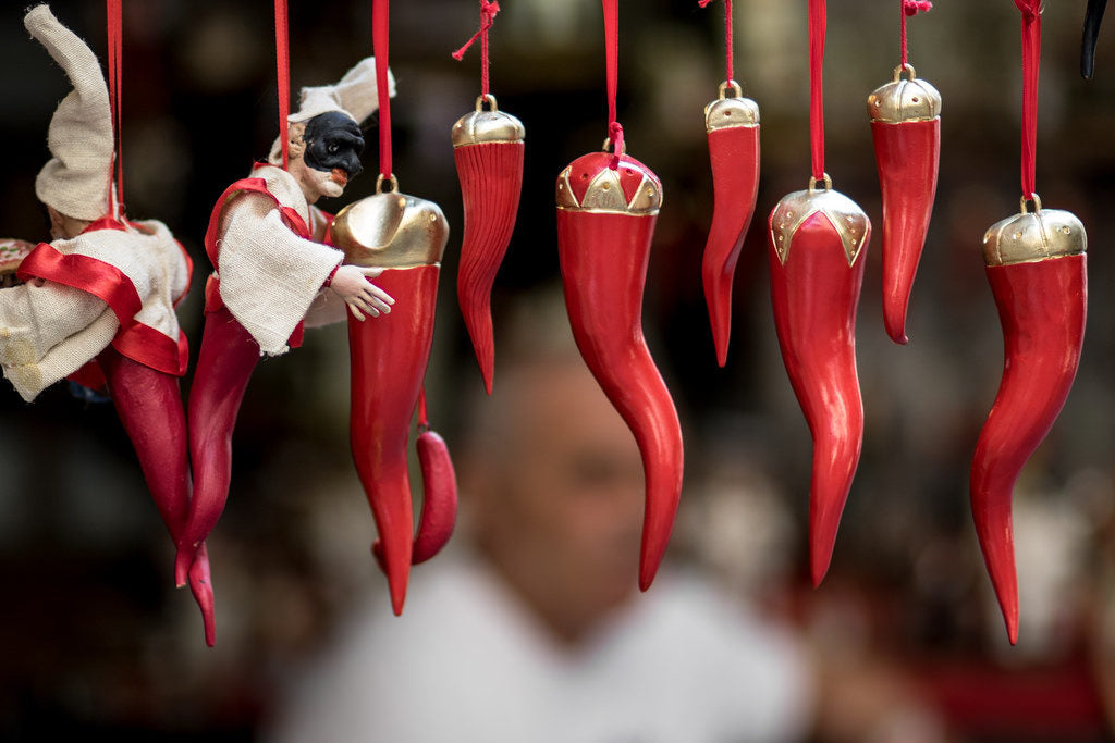 Red horn-shaped amulets and a small doll in a mask hang on red ribbons, set against a blurred background with a person out of focus. The horns have gold caps and are displayed closely together.