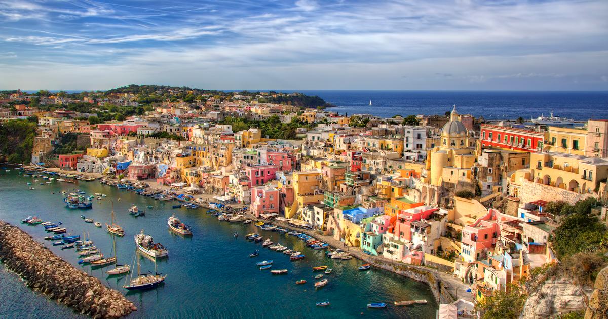A colorful seaside village with pastel buildings on a hillside, small boats docked in a narrow harbor, and a blue sky with wispy clouds above the sparkling sea.