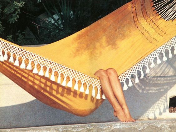 A person relaxes in a yellow hammock with white fringe and tassels, their bare legs and feet extending out, surrounded by sunlight and greenery in the background.