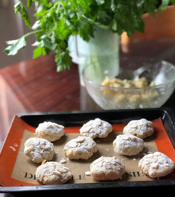 A baking tray with powdered sugar-dusted cookies rests on a table. In the background, a bowl with dough and a vase with leafy green herbs are visible. Sunlight filters into the scene.