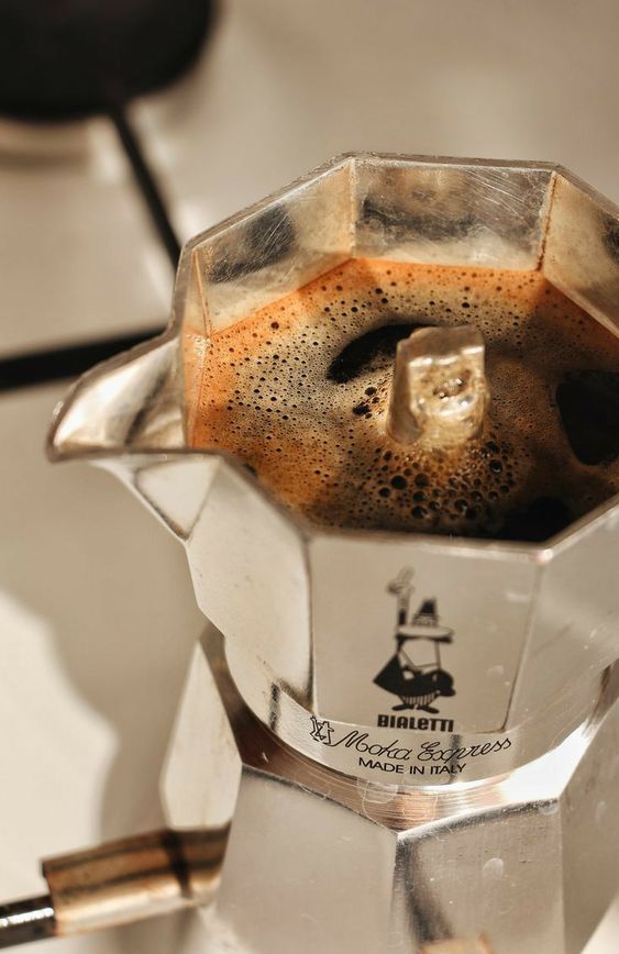 A moka pot brews fresh coffee on a stove, with rich foam visible on the surface. The metallic pot features the Bialetti logo and Made in Italy text.