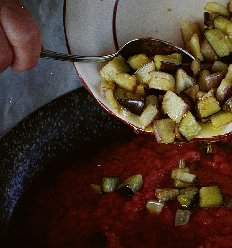 A hand uses a spoon to add cooked diced eggplant from a bowl into a pot of red tomato sauce.