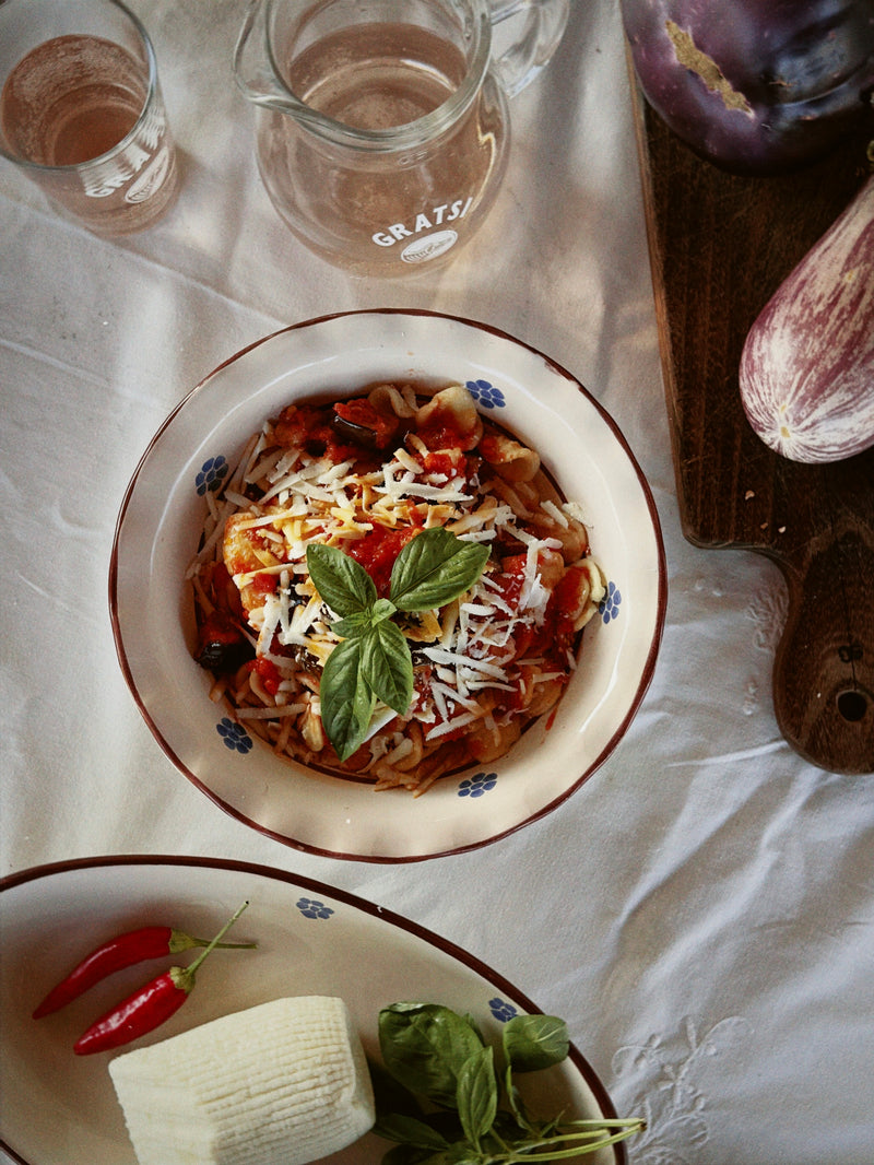 A bowl of pasta topped with tomato sauce, grated cheese, and basil sits on a white tablecloth near a glass pitcher, a block of cheese, red chili peppers, fresh basil, and an eggplant on a wooden board.