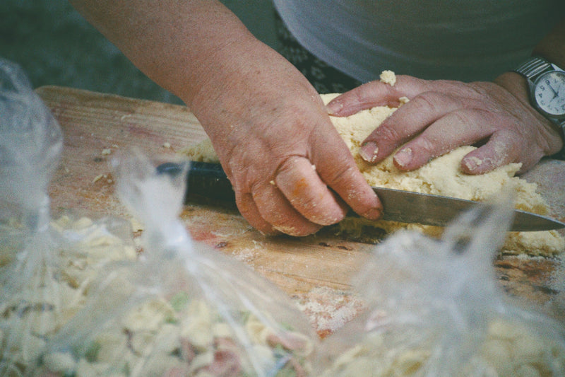 Close-up of a person’s hands cutting dough with a knife on a wooden board. Several plastic bags filled with chopped ingredients are in the foreground. The person is wearing a watch.