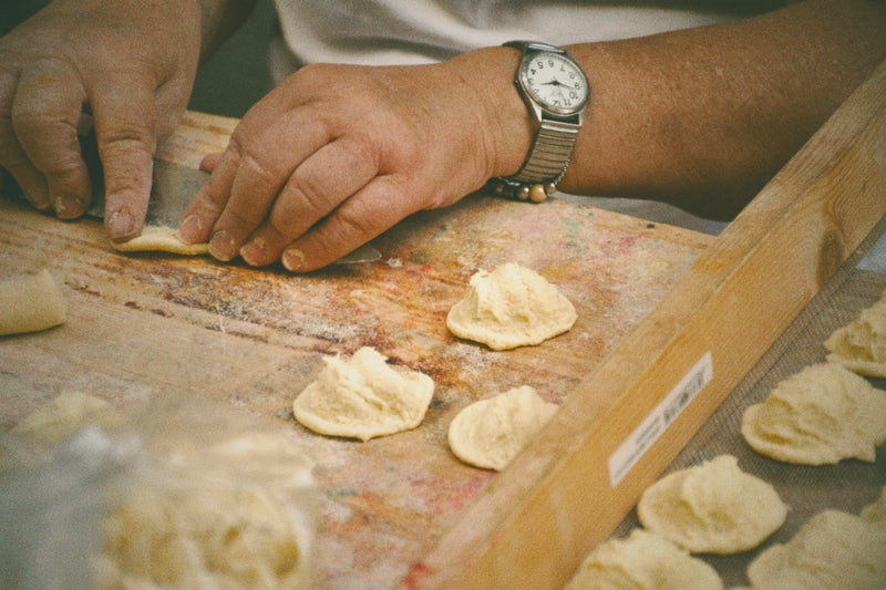 A person wearing a watch shapes dough by hand on a floured wooden surface, with several pieces of dough and small mounds of filling visible.