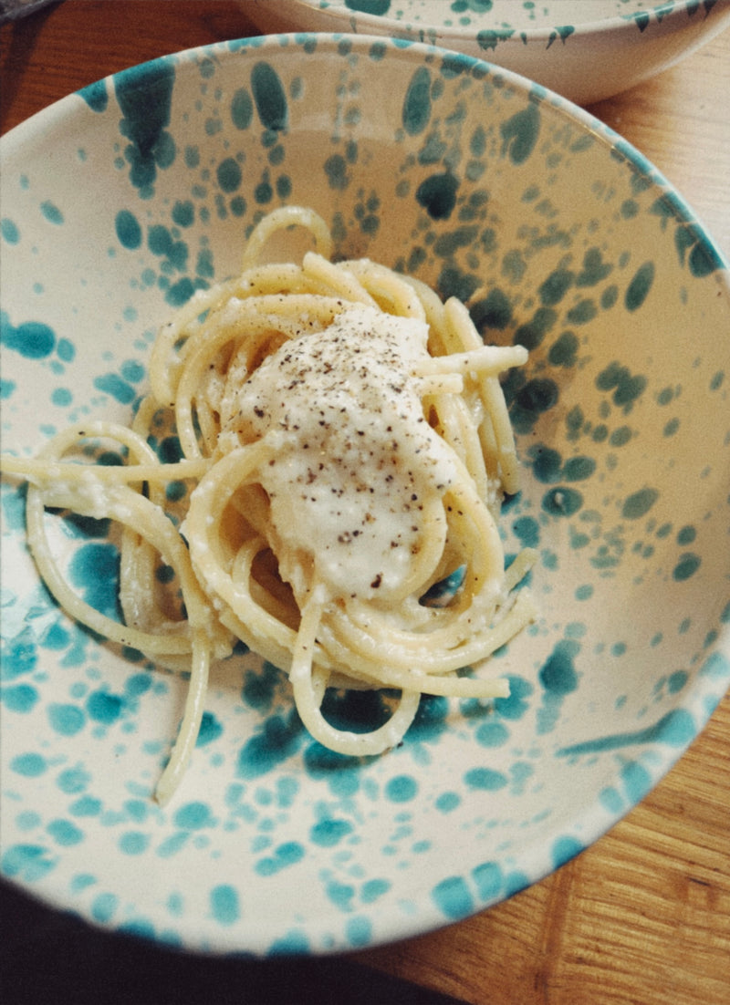 A bowl of spaghetti topped with a creamy white sauce and cracked black pepper, served in a blue and white speckled bowl on a wooden surface.