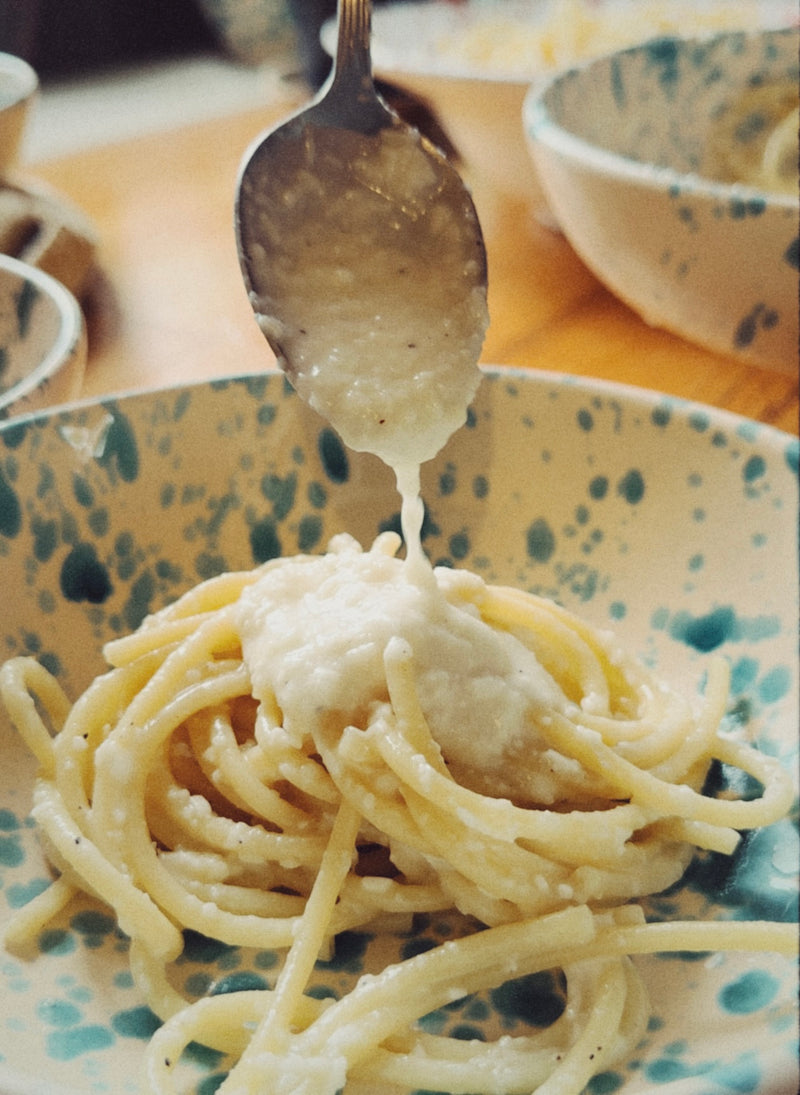 A spoon pours creamy white sauce over a serving of spaghetti in a speckled ceramic bowl on a table.