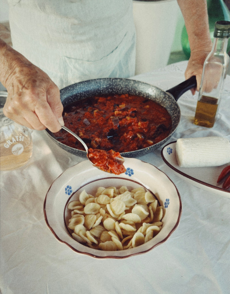 A person spoons red tomato sauce from a pan onto a bowl of shell-shaped pasta, with a bottle of olive oil, a block of cheese, and a glass of grated cheese on a white tablecloth nearby.
